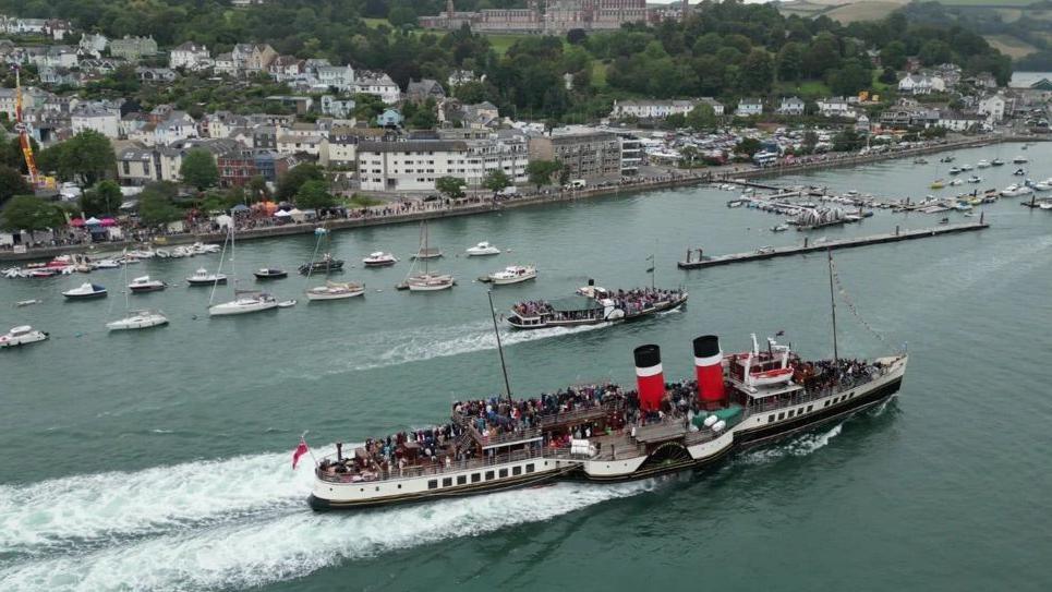 Two paddle steamers, one larger than the other, sailing up the River Dart. There are crowds of people on the boats. There are sailing boats moored in the harbour. 