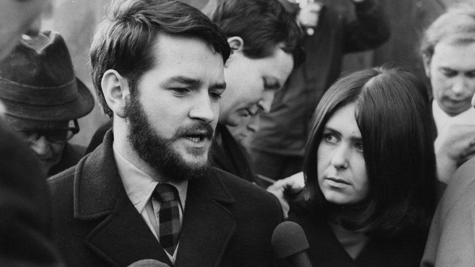 Black and white photo of Dafydd Iwan. He has short dark hair and a beard. He is wearing a shirt and checked tie underneath a dark woollen overcoat. A woman with a long bob hairstyle typical of the 60s and 70s is standing next to him looking at him. He is surrounded by what appear to be reporters writing in notebooks and there are two microphones being held in front of him.