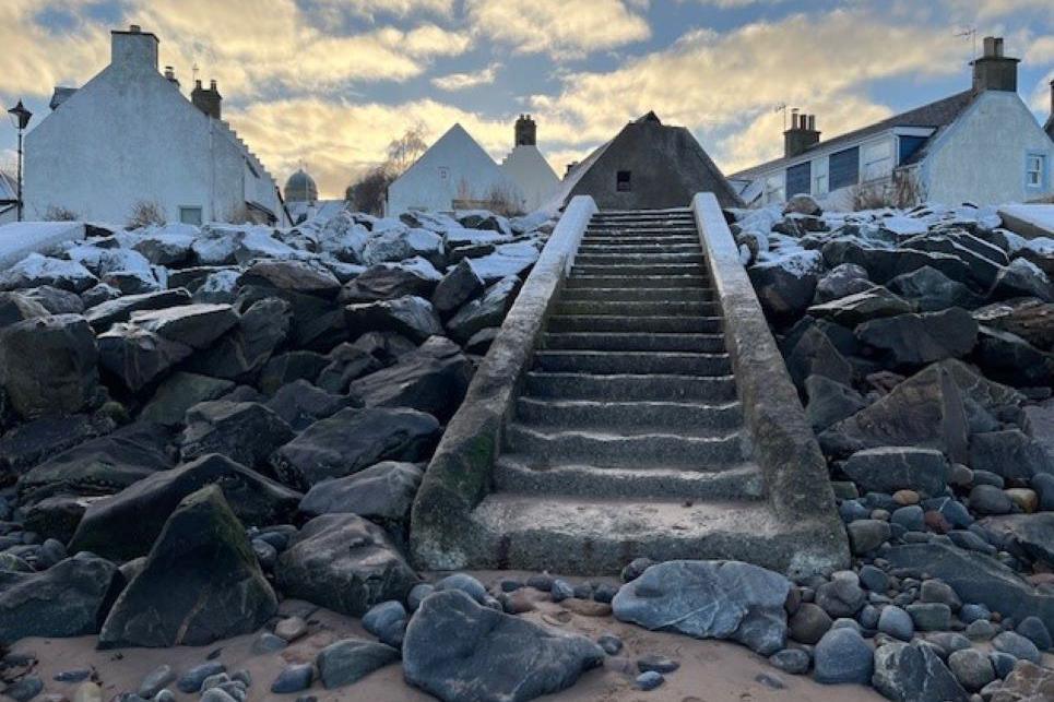 Concrete steps lead from a street down to the beach at Cromarty. Rock armour coastal protection either side of the steps are dusted with snow and there are rows of houses in the background.