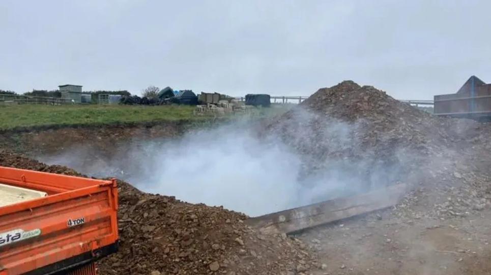 A large square pit cut into the earth is smoking and there is a corner of a truck visible to the left of it.