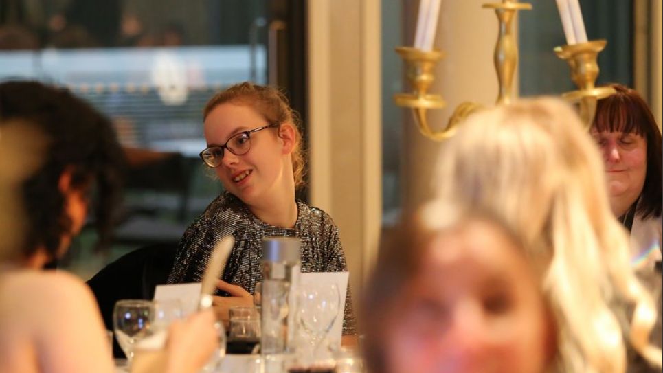 Alisha Burley smiles at she sits at a table in a glamorous-looking ballroom, surrounded by fellow guests