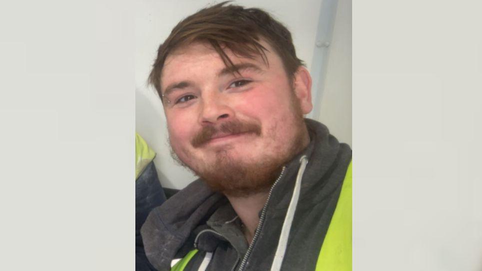 Dean Green is smiling at the camera. He has brown eyes, brown hair and a brown beard and moustache. He is wearing a high-viz jacket over a black hoodie.