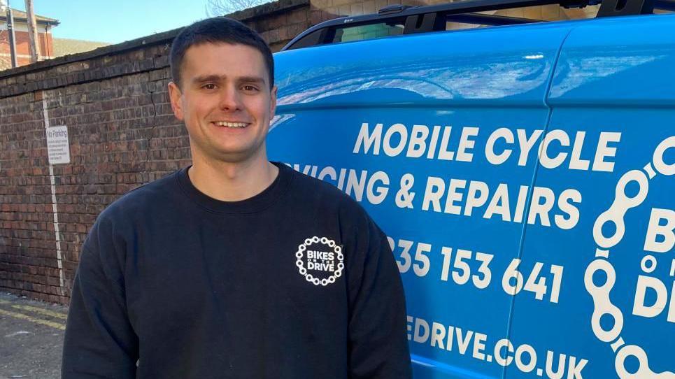 A man in a navy hoodie with a logo that reads "Bikes on the Drive". He is smiling into the camera and has short, dark hair. He is standing in an alleyway with a tall brick wall and is next to a blue van covered in white writing that reads "mobile cycle servicing and repairs". 