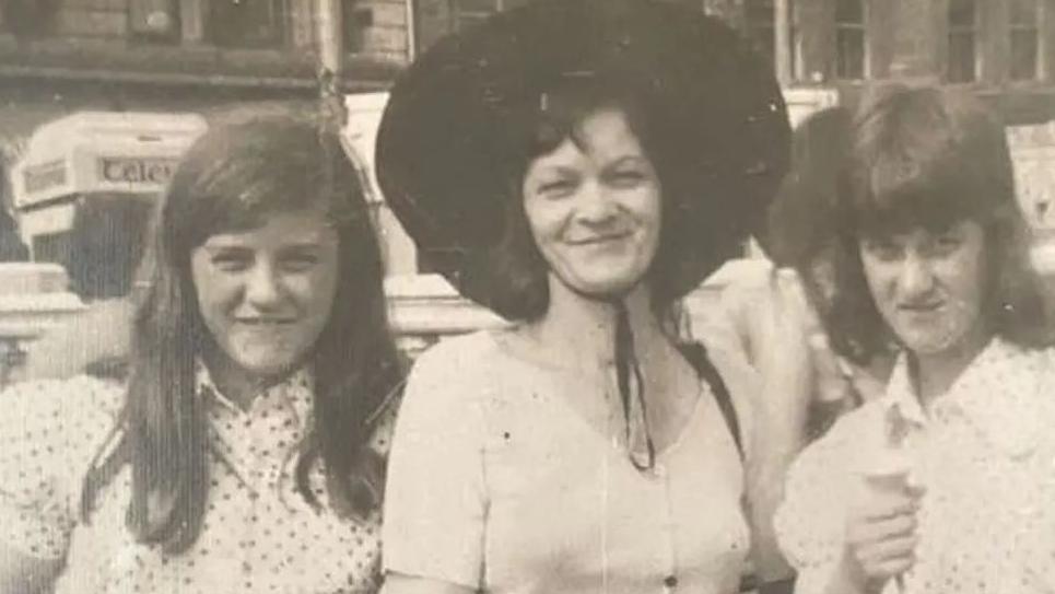 A black and white picture of two girls wearing polka dot shirts standing next to an older woman who is wearing a large black hat tied under her chin.