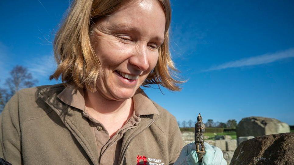 Dr Frances McIntosh who has blonde, short hair holding the knife handle. She's standing in a field and Hadrian's Wall can be seen in the background.