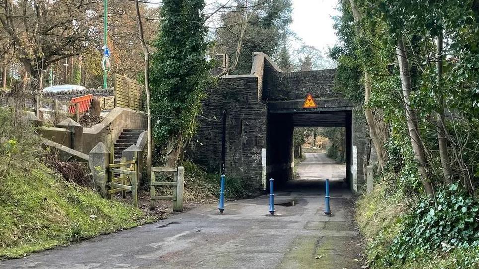 The TT access road, which is lined by trees on the right and has a stone bridge over it. There is set of steps on the left with a gate at the bottom. It runs under a stone-built bridge, which has an yellow height restriction sign on it. There are blue metal bollards in place in front of the tunnel to stop traffic passing through.