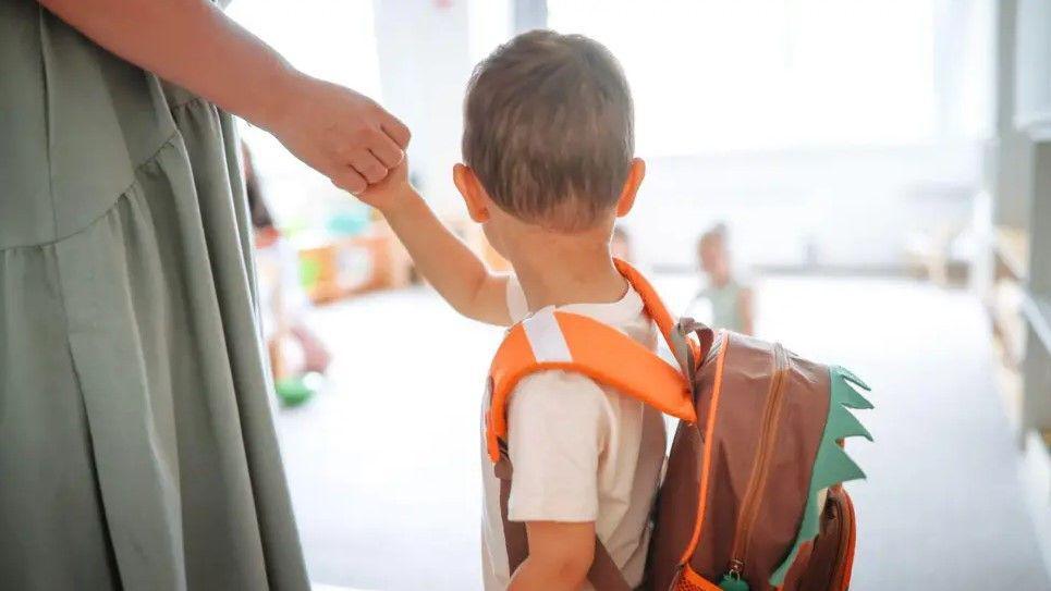A young boy wearing a white t-shirt and an orange and brown rucksack. He is facing away from the camera and a woman in a green dress is holding his hand.