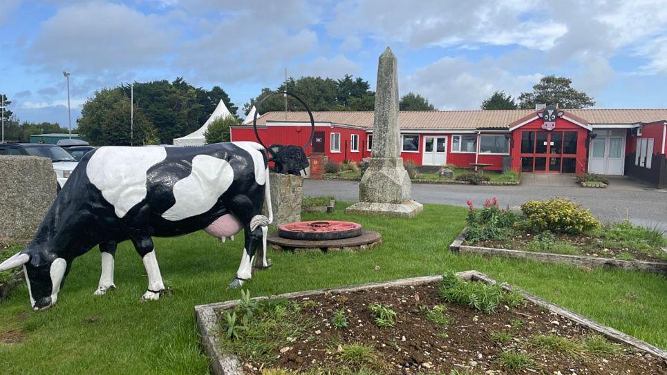 Inside the Dairyland site. In the foreground, there is a life-sized black and white cow statue positioned as if grazing on the grass. Behind the cow, is a small obelisk-shaped stone monument standing upright on a rectangular stone base. In the background is a red building with a beige roof, with large windows and a main entrance with a cow design above the door.