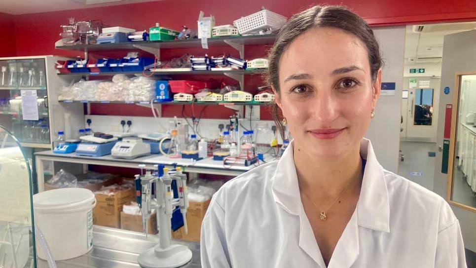 Dr Elizabeth Cooper has tied back brown hair and brown eyes and is slightly smiling. She is wearing a white lab coat and is standing in front of medical testing equipment.