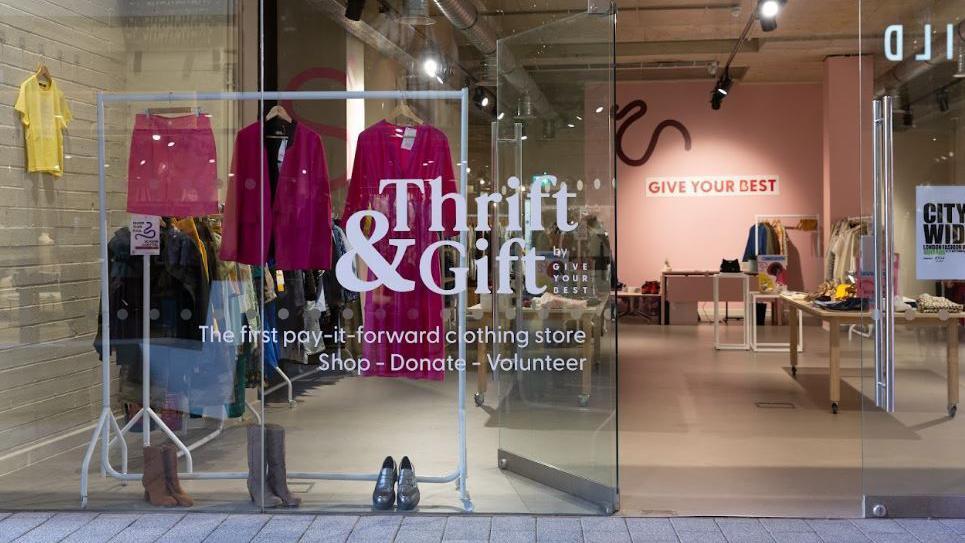 A view of the entrance to the GIve Your Best Thrift & Gift store in London, which has a glass frontage with a white sign on it. Behind the window a white rail can be seen with pink clothes hing above boots and shoes, In the background a pink wall with a red Give Your Best sign on it can be seen, with tables and rails of clothes dotted around