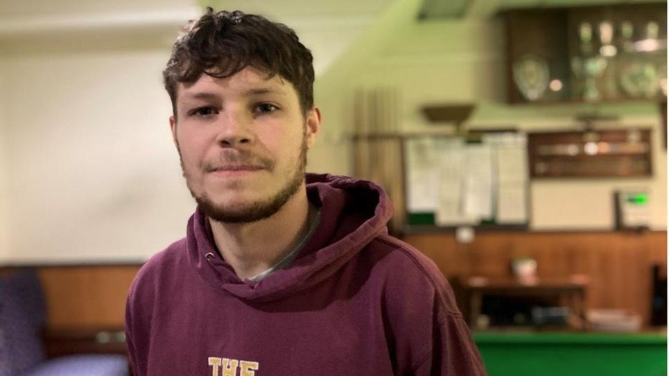 A young man wearing a purple hoodie stands in a snooker room in front of a table and trophy cabinet. He has short, curly brown hair with a fringe, and a neatly trimmed brown beard.