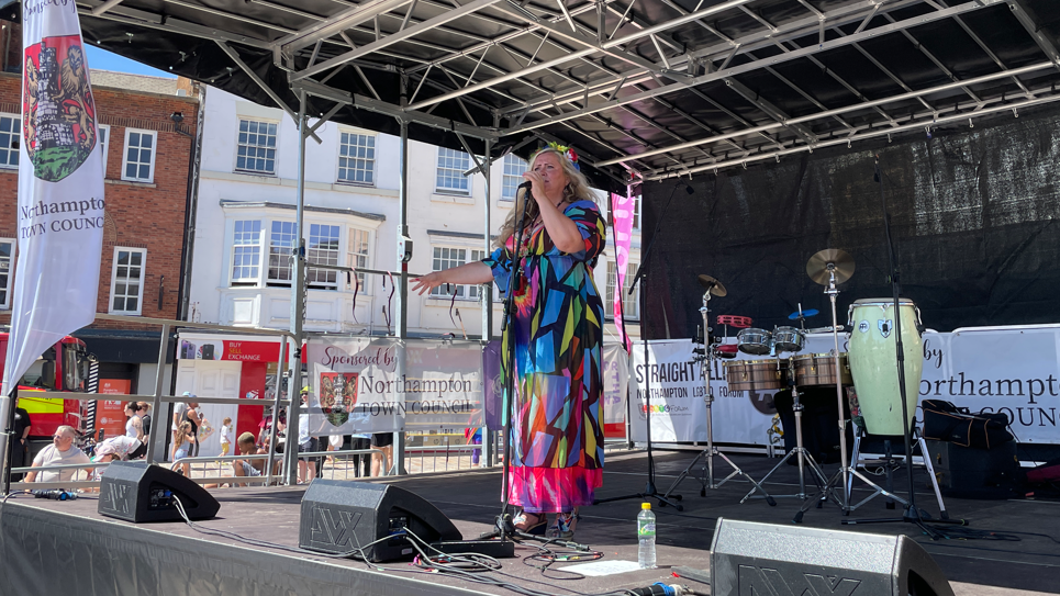 A woman is standing on a stage and singing into a microphone. She is wearing a multi-coloured long dress.