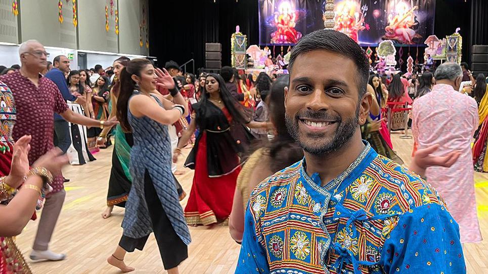 Hiran Patel wearing a blue outfit smiling as people dance as part of the Navratri festival 