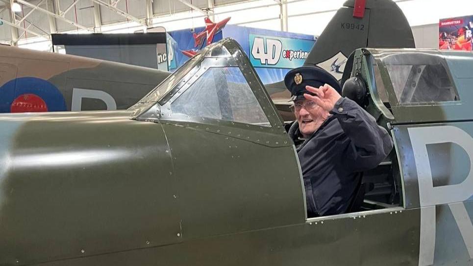 An elderly man is sitting in the cockpit of a Spitfire, wearing a peaked cap and saluting while he looks at the camera