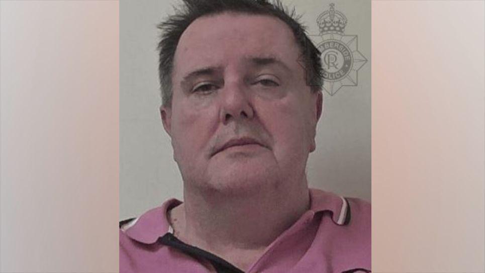 A police head and shoulders shot of a man with brown hair with some of the front sticking out. He is pulling a serious expression and wearing a pink polo shirt.