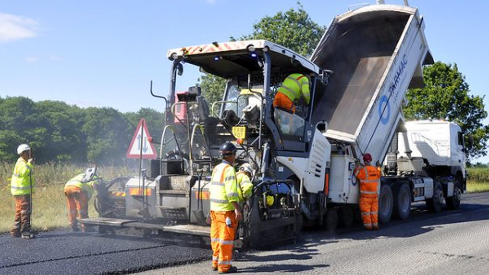 A large white van is tipping concrete onto a road, which is black in colour and steaming. There are five workers visible, who are in orange and yellow hi-vis trousers and jackets.