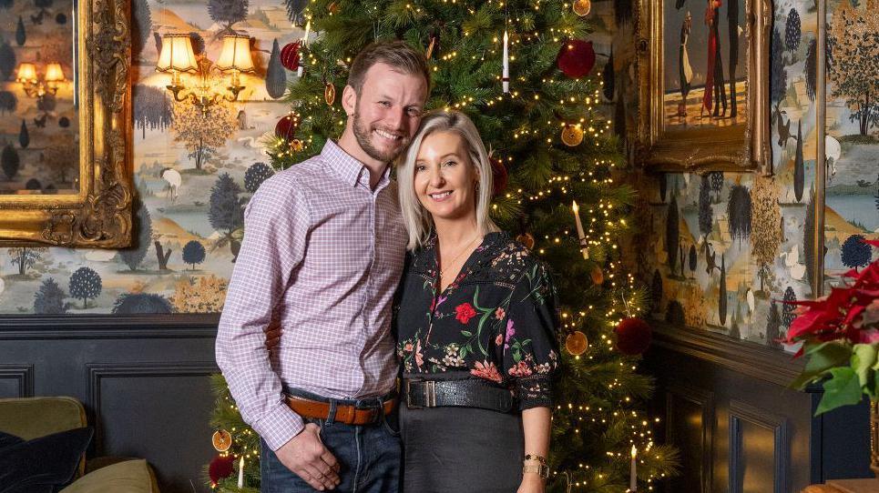 A man and woman standing smiling in front of a Christmas tree.