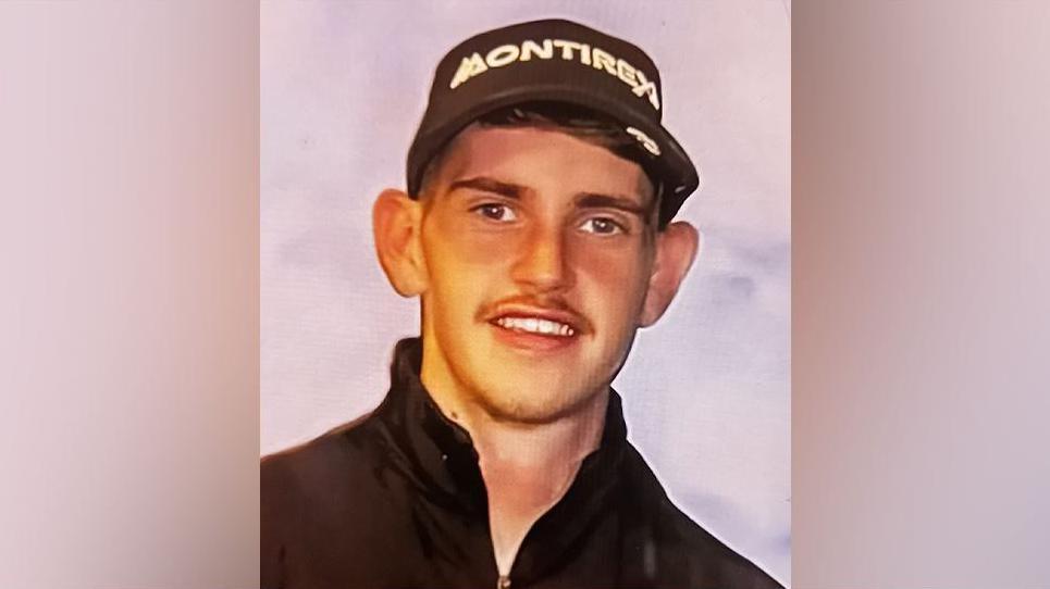 Head and shoulder shot of a young man with short dark hair and a faint moustache. He is half-smiling and wearing a black fleece or jacket and a black cap with white letting spelling out Montirex