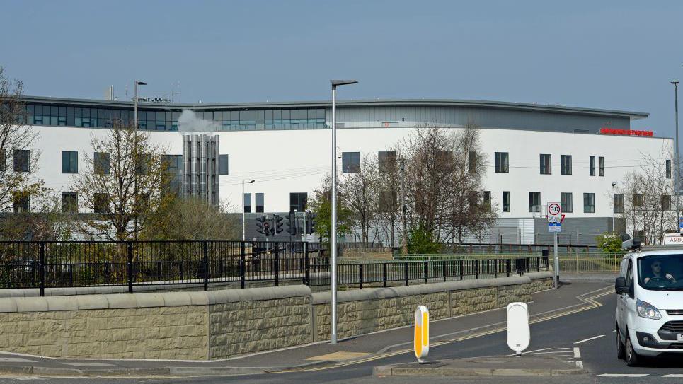 A white hospital building behind a low brick wall and black gate with street lamps and a road around it