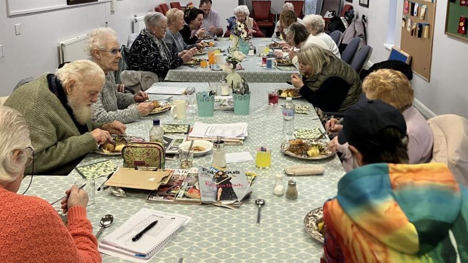 A large group of older people are sat round two tables, with green and while table cloths, eating lunch and socialising at their weekly lunch club.