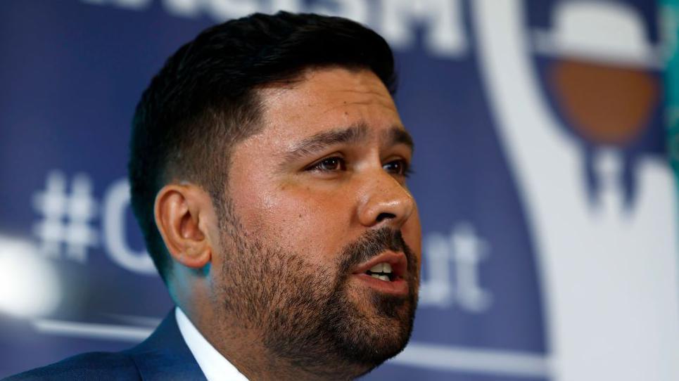 Former Scotland player Qasim Sheikh in close up with a Running out Racism board behind him. He has a beard and a white collared shirt with a dark blue jacket on.