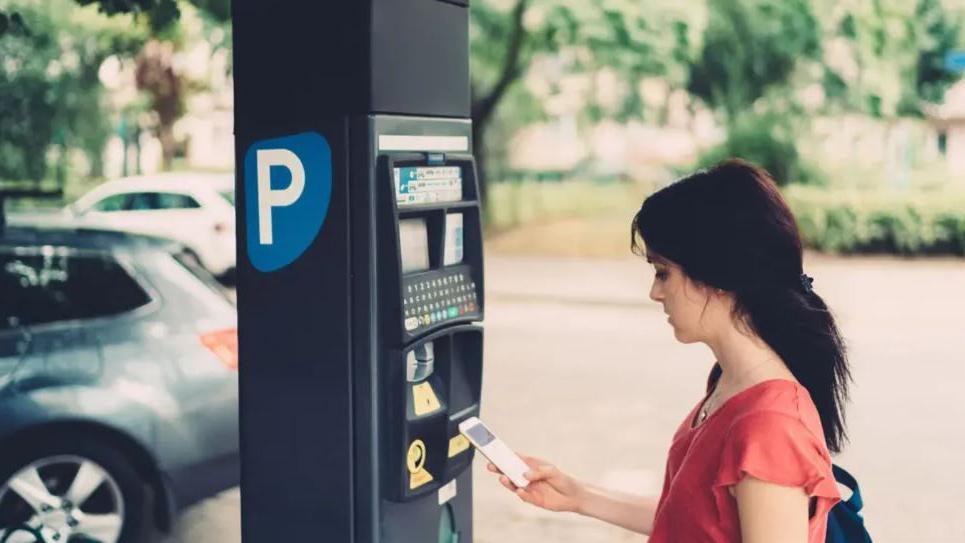 A woman looks down at her mobile phone as she starts next to a parking payment machine