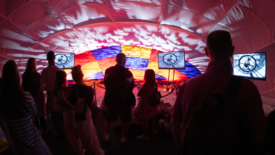 Eight people are standing with their backs to the camera. They appear to be inside a hot air balloon canopy. Lights are being projected onto the canopy and the people are silhouetted. There are three TV screens simulating what it is like to fly inside a balloon.