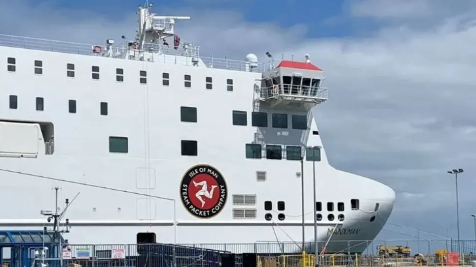 The bow of the Manxman ferry moored in Douglas Harbour. It is a white vessel with the Isle of Man Steam Packet Company's logo on the side, which features a white three-legs-of-Mann symbol in a red circle with a black band around it featuring the company's name in white lettering.