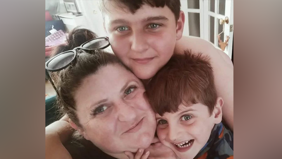 A woman and two young boys huddle together and smile into the camera. The woman has dark hair pulled back from her face and dark framed glasses perched on her head. The two boys have brown hair.