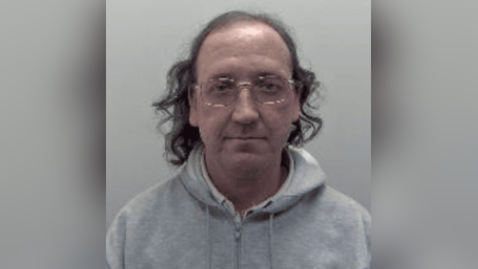 Police mugshot of a balding man with straggly long black hair and glasses. He is wearing a grey hoodie.
