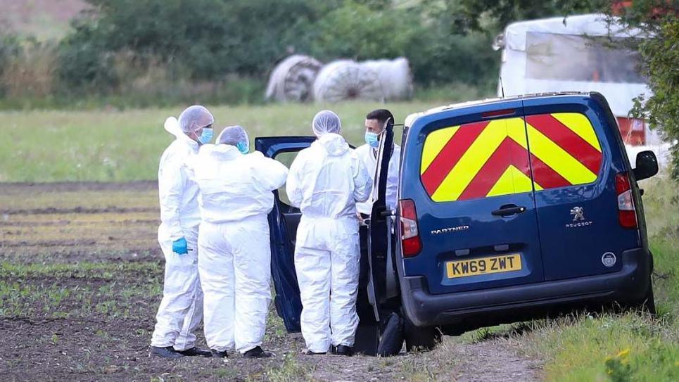 Four forensic officers, dressed in white suits and wearing blue masks and white hairnets, stand next to a blue van in a field