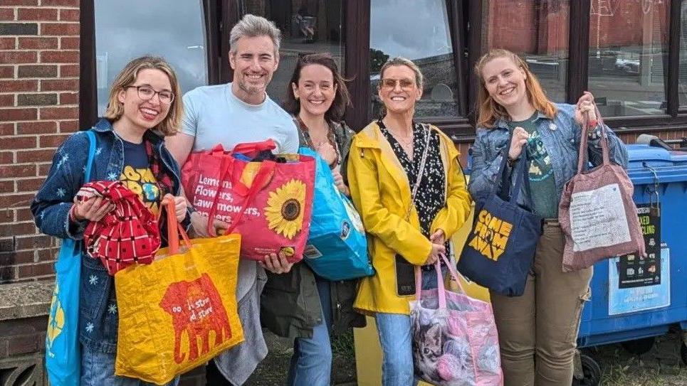 Four women and a man are smiling whilst posing for the camera and holding large bags which appear to contain charitable donations.