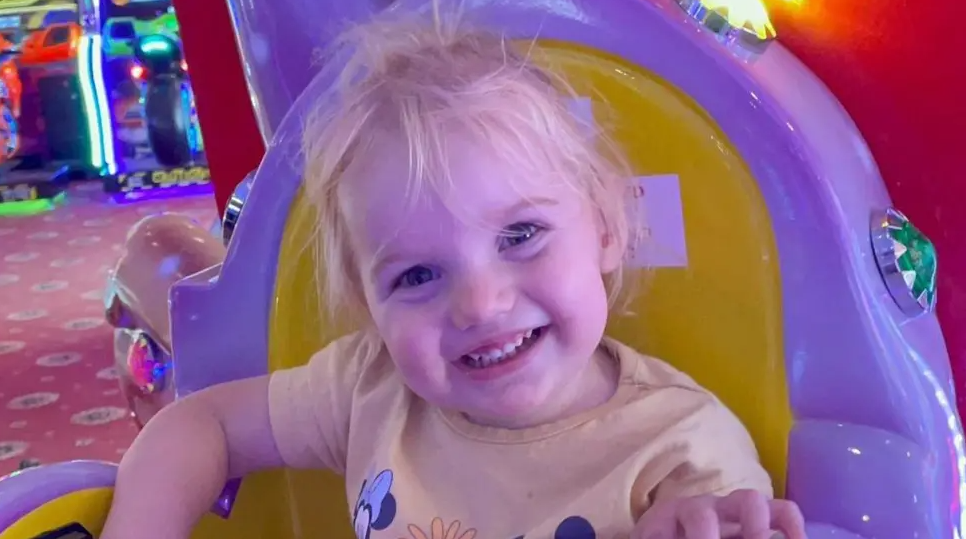 A young blonde toddler wearing a yellow t shirt smiling into the camera. She is sitting on a yellow and purple chair at an arcade.
