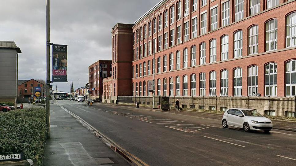 Part of the route where a city-bound bus lane has been approved showing a wide road with double yellow lines outside a supermarket which is opposite a large red brick mill with lots of windows. A small white car is driving down the road to the right hand side of the image