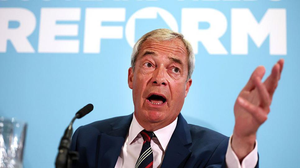 Nigel Farage, dressed in a dark suit, sits on a podium before a microphone with the words Reform written on a blue background behind him