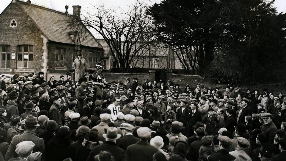 A black-and-white archive photograph from 1938 showing a large crowd gathered around the "fool" ahead of the game.