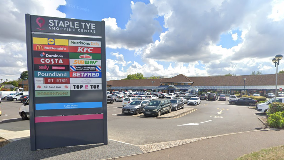 The car park at Staple Tye Shopping Centre in Harlow. On the left is a grey sign which reads "Staple Tye Shopping Centre", and the sign lists the shops and restaurants at the centre