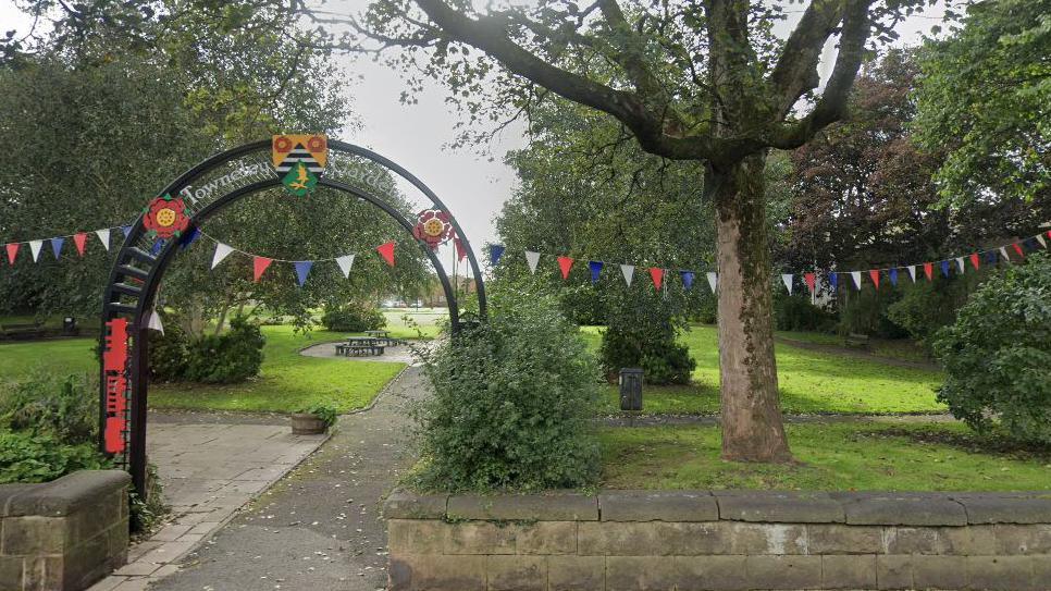 Street view image of the entrance to Towneley Gardens, showing a black metal archway saying Towneley Gardens, flat grassed areas and established trees, strung with red white and blue bunting