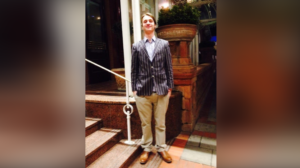 A young man in a pin strip blazer and cream chinos smiles standing straight.