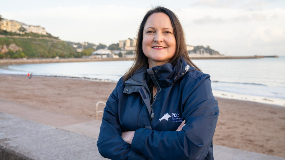 A woman with her arms crossed, stood on a beach looking towards the camera. She is wearing a navy blue waterproof coat and has brown hair.