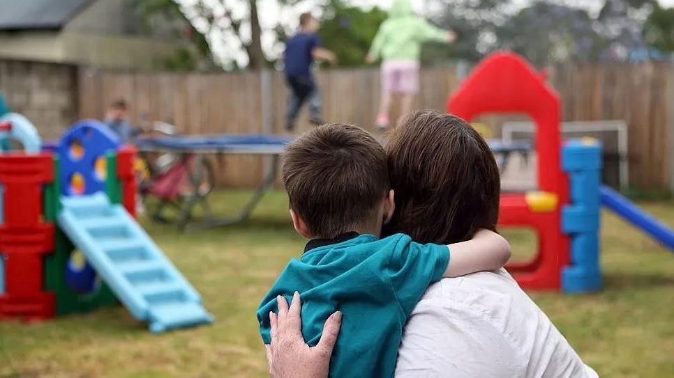 A woman holding a young boy in a park, their faces turned away