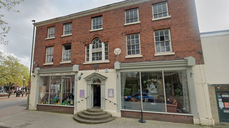 A large building housing two shops. The top half is red brick and the bottom half has large windows wither side of a door. There are stone steps leading to the door, which is green and cream and has black hand rails.