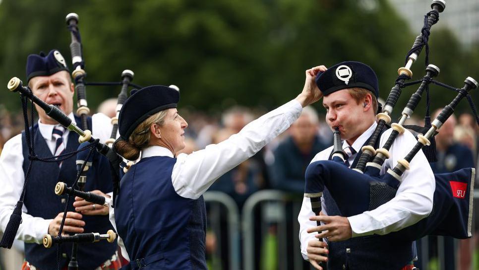 Female pipers fixes the hat of a male pipers as he blows in the bagpipes.