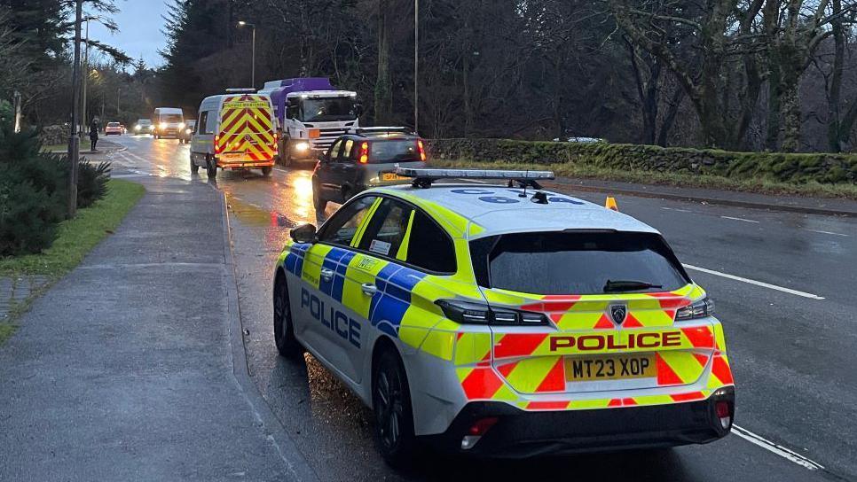 A police car is parked in a small layby on a busy road. There are cars, vans and a lorry with their lights on on the road up ahead.