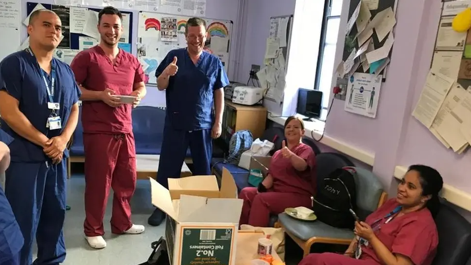 Five hospital workers congregated around an open cardboard box. Two are wearing blue scrubs, and three are wearing mauve scrubs.