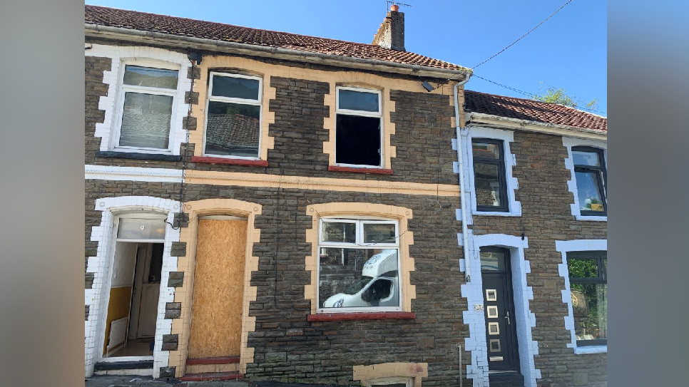 The outside of a terraced house with a boarded up doorway and a broken window