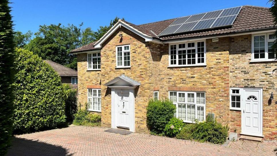 A five-bed detached home. It has a hedge in front of the building and on the side of the property. The property also has solar panels installed on the roof.