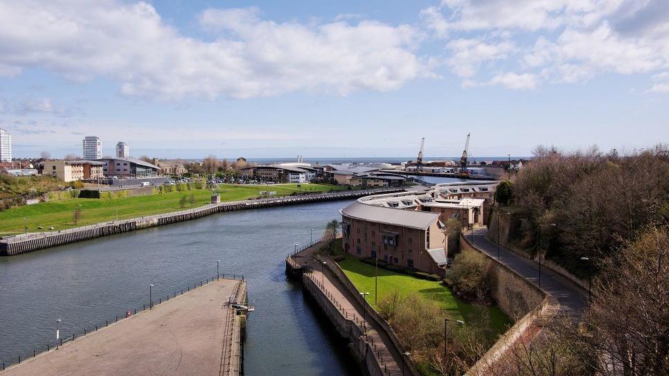 A view of the River Wear with large modern buildings on either side. On the left is a large patch of grass. Further down the river there are two large cranes. The sea can be seen beyond them.