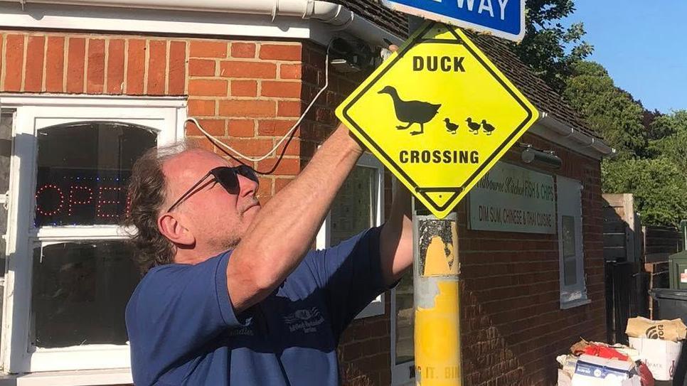 A man wearing a blue t-shirt and sunglasses, attaching a yellow diamond-shaped sign to a lamppost using cable ties. The sign says 'duck crossing', with a silhouette image of a mother duck being followed by three ducklings.