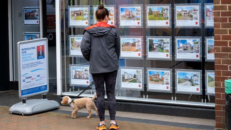 Woman in running gear and a coat, with a small dog on a lead, looks at adverts in an estate agents window.